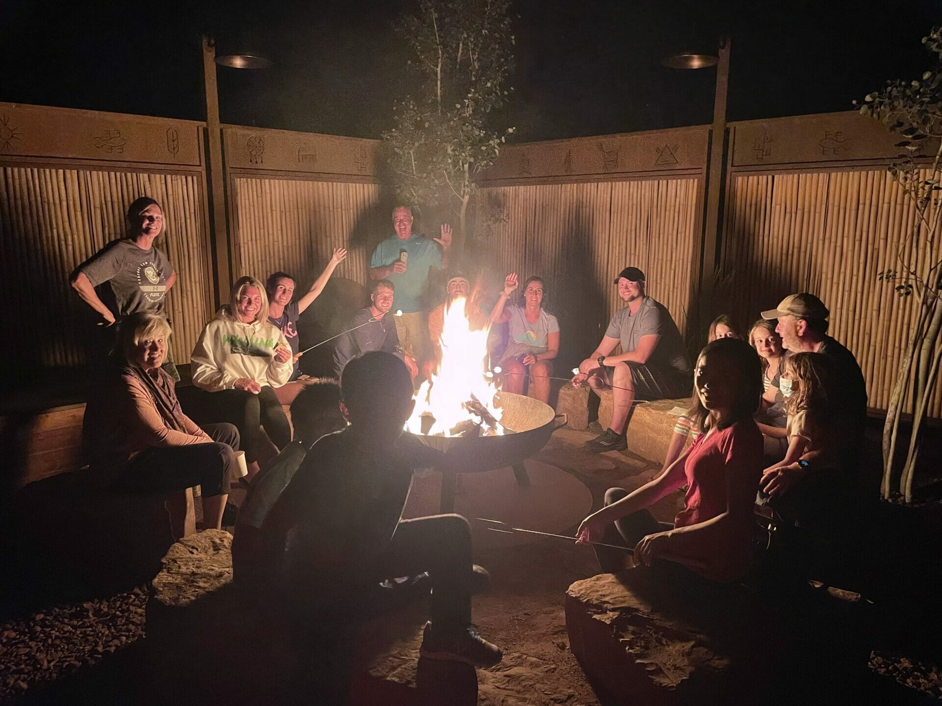 several families sit around a roaring bonfire, a wooden fence behind them.