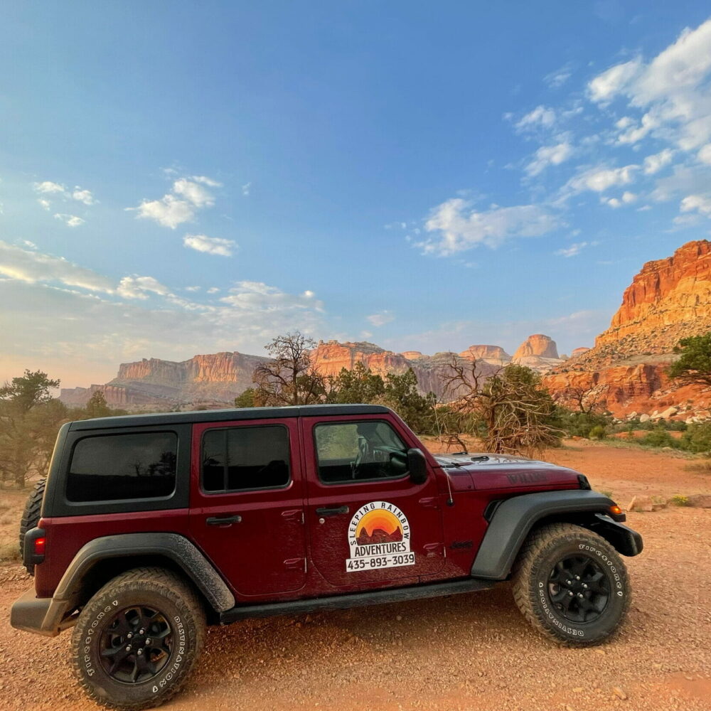 Dark red sleeping rainbow adventures jeep parked on dirt road, the capitol reef scenic drive is in the background