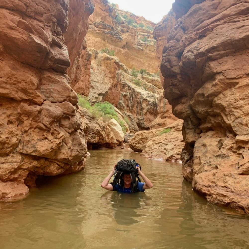 hiker in water up to his neck inside a Sulphur Creek, Capitol Reef National Park. He's holding his bag over his head